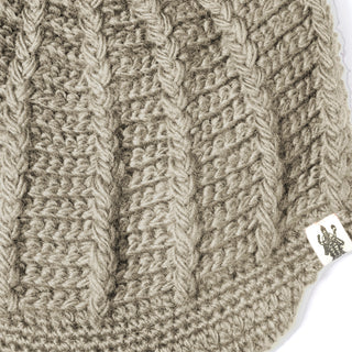 Close-up of the Hand Crochet Martha Cap in beige, highlighting its intricate braided knit pattern and a small white tag on the bottom right with an illustration of a person holding an object—an ideal display of the exceptional craftsmanship that defines this handmade wool hat.