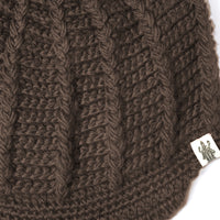 Close-up of the Hand Crochet Martha Cap, showcasing its brown knitted fabric with a ribbed pattern evocative of handcrafted wool. A small white tag featuring a black illustration is sewn onto the lower edge.