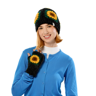 A person with a smile is wearing sunflowers-adorned black knit hat along with matching Sunflower Handwarmers. They are also wearing a blue button-up sweater over a white shirt. The background is plain and black.