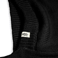 A close-up view of the black Hand Loom Nomad Hood Scarf, featuring a small white tag sewn onto the material with a simple animal illustration, possibly a lizard or crocodile. The background is white.