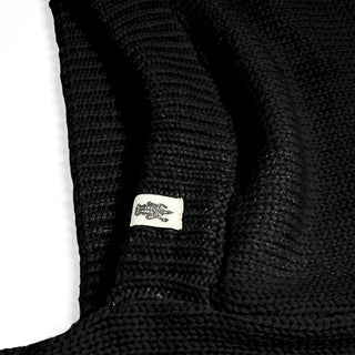 A close-up view of the black Hand Loom Nomad Hood Scarf, featuring a small white tag sewn onto the material with a simple animal illustration, possibly a lizard or crocodile. The background is white.