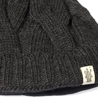 A close-up of the Hand Knit Nord Hat, a dark beanie with a ribbed cuff and a small beige fabric tag adorned with a simple black illustrated design. This hat is lined with cozy fleece for extra warmth and comfort.
