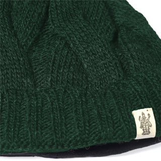 Close-up of the Hand Knit Nord Hat—this dark green beanie features a visible ribbed pattern and a small beige fabric tag near the edge, adorned with a black illustration of a figure.