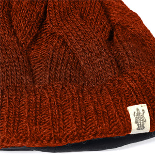 A close-up of the Hand Knit Nord Hat, a rust-colored beanie with a cable pattern and cuffed edge. This hat is crafted from hand knit wool and features a small sewn-on tag with a black printed graphic near the ribbed brim.