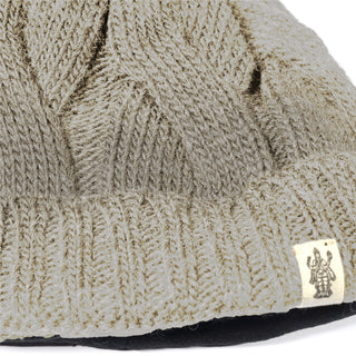 A close-up of the Hand Knit Nord Hat, a beige cuffed beanie with a textured cable pattern, hand knit from wool and featuring a small tag sewn onto the folded brim with a black graphic of two robed figures.
