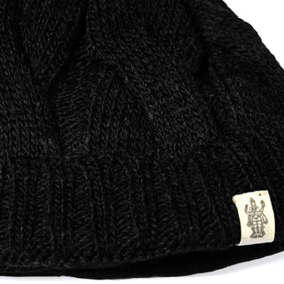 Close-up of the Hand Knit Nord Hat, a black hand-knit wool beanie featuring a distinct ribbed pattern and a small off-white fabric tag on the edge with a dark printed figure graphic.