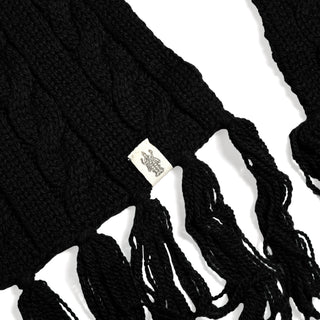 Close-up of the Hand Knit Plush Scarf in black, crafted from soft Merino wool with prominent thick cable patterns and large fringe ends. A small white fabric tag with a dark design accents one end, and the scarf is set against a clean white background.