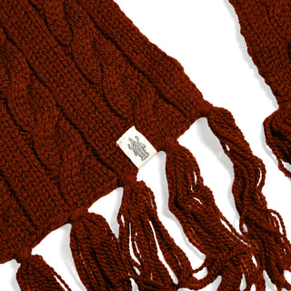 A close-up of the Hand Knit Plush Scarf showcases its thick, dark red Merino wool construction and classic cable pattern. The scarf features fringe ends and a small white fabric tag with a black graphic logo sewn onto it. The background is white.