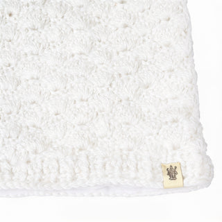 A close-up of the Hand Knit Puff Beanie showcases its white, textured knit fabric, reminiscent of handcrafted work. A small beige tag with a brown design is sewn onto the bottom edge, set against a plain white background.