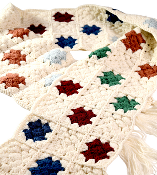 The Crochet Patch Scarf showcases a vibrant pattern of diamond shapes in an array of colors—blue, green, red, brown, and light blue—interspersed with white sections. The scarf is displayed to highlight its length and striking colorful design.