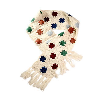 The Crochet Patch Scarf is a long, cream-colored piece with tasseled ends, showcasing a vibrant pattern of multicolored star-shaped designs in blue, green, red, and brown set against a black background.