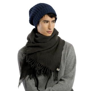 A person wearing a dark gray Merino Fringe Scarf, handmade in Nepal, draped around their neck and shoulders, coupled with a blue beanie hat. The individual is clad in a gray textured shirt and is glancing slightly to the side against a black background.