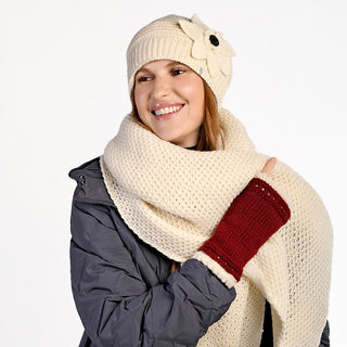 Woman smiling and looking to the side against a plain white background, wearing a gray jacket, cream knit hat with a flower, red fingerless gloves, and the handmade Ithaca Scarf from Nepal—a cheerful winter accessory ensemble.