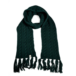 The Hand Knit Plush Scarf is a dark green, hand-knit cable scarf made from soft Merino wool. It features long fringe on both ends and has a small white tag near one end, shown against a white background.