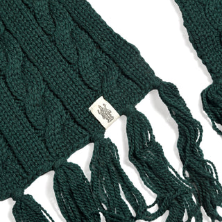 A close-up of the Hand Knit Plush Scarf in dark green, featuring cable stitching and fringe ends. A small white fabric tag with a logo is attached to the Merino wool scarf, which is laid flat on a white background.