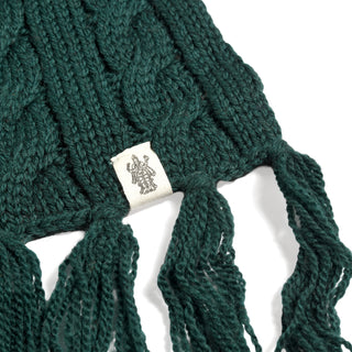 A close-up of the Hand Knit Plush Scarf in dark green, crafted from soft Merino wool with cable detailing, finished with fringed ends and a white fabric tag displaying an illustration of two figures in medieval-style clothing.