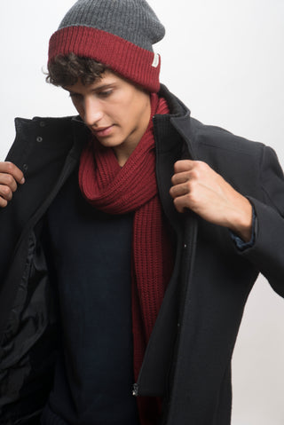 A man in a dark coat, wearing a Hand Loom Laurent Scarf handmade in Nepal and a gray-red knit beanie, looks down while adjusting his coat against a plain light background.