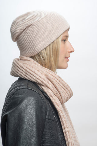 A person in profile wears a light pink Hand Loom Laurent Scarf and matching knit beanie with a black leather jacket. The merino wool scarf is handmade in Nepal. Plain white background; straight blonde hair.
