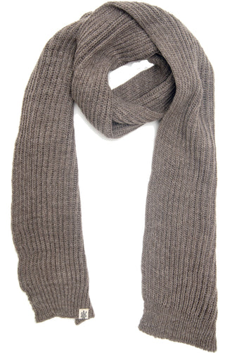 The Laurent Scarf is a long, thick ribbed-knit merino wool scarf in light brown, handmade in Nepal. It’s shown laid flat with one end looped into a circle and features a small fabric tag near the end.