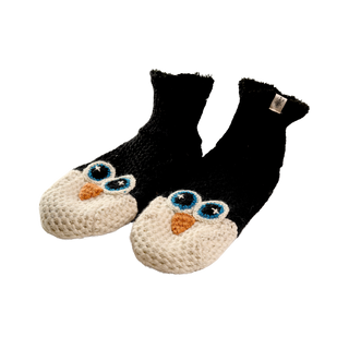 The Penguin Slippers are a pair of black knitted slippers with white tips, designed to resemble penguin faces. They feature blue eyes and orange beaks and are hand-knit in Nepal from 100% wool, giving them a soft and cozy appearance perfect for indoor use.