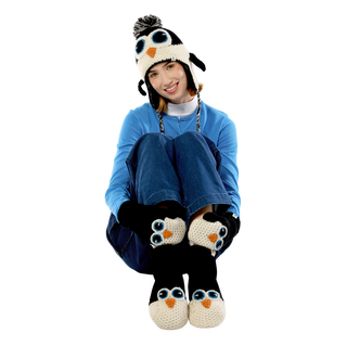 A person in a blue outfit is sitting with knees up and arms wrapped around them. They are wearing a knitted penguin hat, matching mittens, and Penguin Slippers made from 100% wool, hand-knit in Nepal. The ensemble features large blue eyes and beak decorations, resembling penguins.