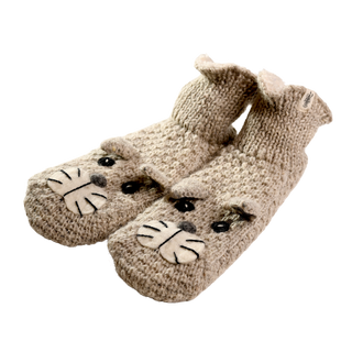 Introducing Shmil The Cat Slippers: A pair of beige, knitted baby booties with adorable cat faces on the toes, lovingly hand-knit in Nepal. These cozy slippers feature black eyes, a white nose, and whiskers on the cat faces. Crafted from soft merino wool, they have ribbed cuffs for a snug fit against the plain black background.