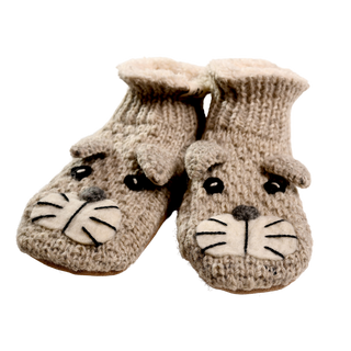 Shmil The Cat Slippers are hand-knit in Nepal from soft merino wool. These beige slippers showcase adorable animal faces with black button eyes, white felt snouts, and dark stitched whiskers. They feature a ribbed pattern on top and have a soft, fluffy interior for enhanced comfort.