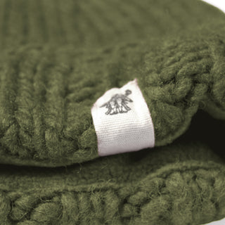 A close-up of thick, green knitted fabric from the Hand Knit Timber Hat shows a white tag sewn onto it, featuring a small black graphic or logo.