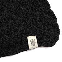 A black Hand Crochet Veil Scarf, crafted in a textured bobble stitch from Merino wool, featuring a small white label sewn at the edge with a line drawing of a multi-armed figure.