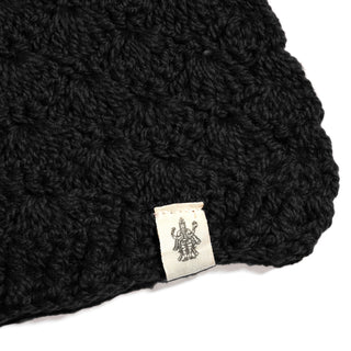 A black Hand Crochet Veil Scarf, crafted in a textured bobble stitch from Merino wool, featuring a small white label sewn at the edge with a line drawing of a multi-armed figure.