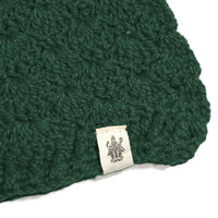 A close-up of the Hand Crochet Veil Scarf, crafted from dark green yarn in a textured bobble stitch, with a cream-colored label attached that displays a black line drawing of a multi-armed figure.