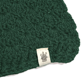 A close-up of the Hand Crochet Veil Scarf, crafted from dark green yarn in a textured bobble stitch, with a cream-colored label attached that displays a black line drawing of a multi-armed figure.