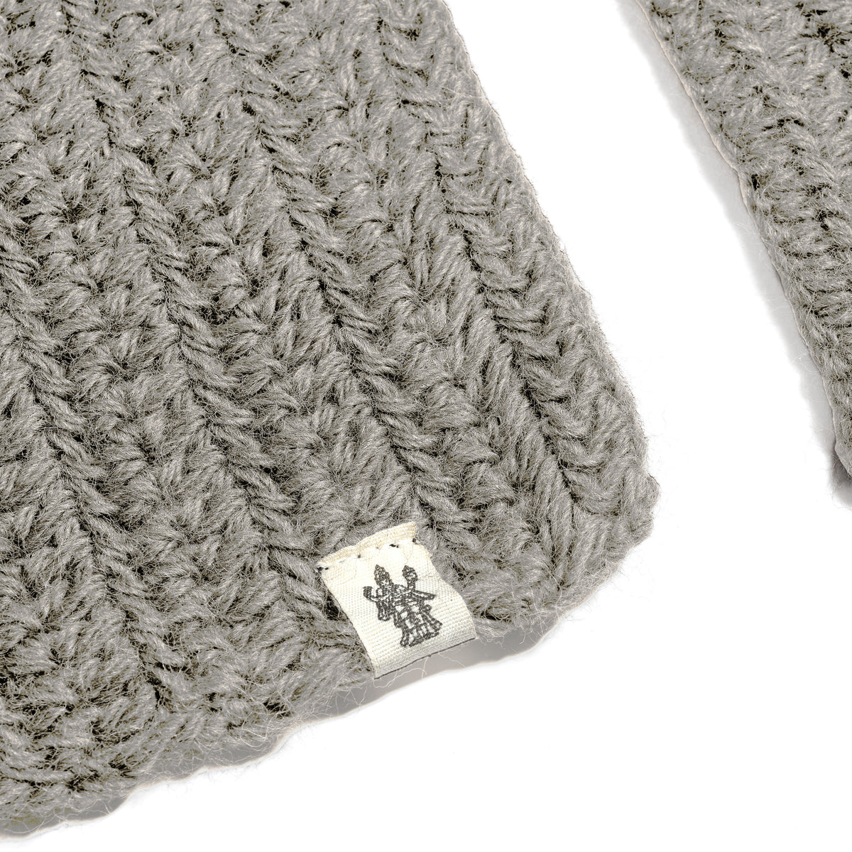 A close-up of a gray knitted fabric with a small white tag featuring a dark symbol resembling a trident. The ribbed crochet texture is clearly visible, giving the Hand Crochet Velour Scarf a distinctive and cozy appearance.