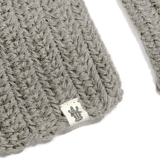 A close-up of a gray knitted fabric with a small white tag featuring a dark symbol resembling a trident. The ribbed crochet texture is clearly visible, giving the Hand Crochet Velour Scarf a distinctive and cozy appearance.