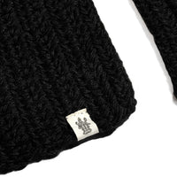 A close-up of the Hand Crochet Velour Scarf in black, showcasing its ribbed crochet texture with a small white label featuring a decorative logo sewn onto the edge. The background is plain white.