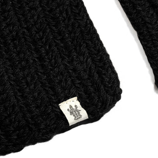 A close-up of the Hand Crochet Velour Scarf in black, showcasing its ribbed crochet texture with a small white label featuring a decorative logo sewn onto the edge. The background is plain white.