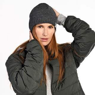 A person with long auburn hair wears the Dakota Hat in gray wool, a gray sweatshirt, and a dark green puffer jacket. One hand rests on their head, the other touches their ear as they face the camera with a neutral expression.