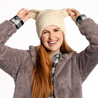 A person with long red hair smiles while holding the pointed tips of a cream Bear Ear Beanie. They're dressed in a gray fleece jacket over a plaid shirt, standing against a plain white background.