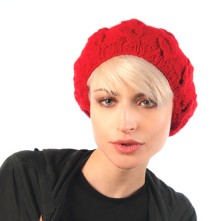 Woman wearing a red Cable Beret hat handmade in Nepal with a braid knit design and black top, with short blonde hair, looking directly at the camera.