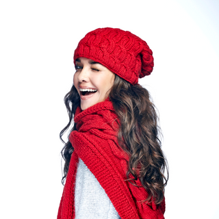 A woman with long wavy hair smiling and looking over her shoulder, wearing a red handmade Nepal knitted hat and a Lucky Knit Scarf with a horseshoe rib knit design.