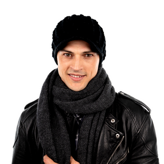 A man wearing a black leather jacket, a handmade Knit Jockey Cap, and a scarf, smiling at the camera against a white background.