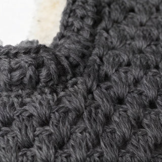 Close-up of the Hand Crochet Cloud Hood in dark gray, highlighting its thick yarn and interlocking stitches. A small section of off-white material is visible in the corner.