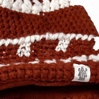 A close-up of the Hand Crochet Discover Hat reveals its textured, chunky knit, crafted from soft lambswool in brown and white with an alternating yarn color pattern. A small tag featuring a black line drawing of a figure is attached to the fabric.