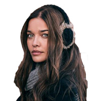 A woman with long brown hair, wearing Hand Knit Cable Adjustable Earmuffs With Faux Fur, along with a gray scarf and a dark coat, looks toward the camera against a white background.