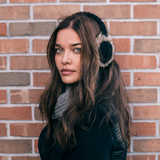 A woman wearing Cable Knit Adjustable Earmuffs with faux fur in front of a brick wall is holding a device with wireless charging.