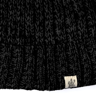 Close-up of the Hand Knit Grid hat, a black Merino wool beanie with a prominent ribbed texture. A small beige fabric tag displaying a graphic of a person holding an axe is sewn onto the lower edge.