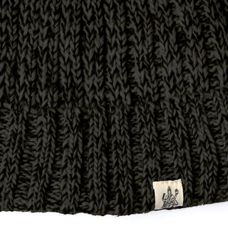 Close-up of a black knitted fabric from the Hand Knit Grid hat, featuring a small beige tag with a simple line drawing of a seated figure, possibly holding an object.
