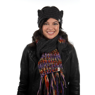 A woman is smiling at the camera, wearing a black leather jacket, a Kit Kat headband, and a multicolored knitted scarf.