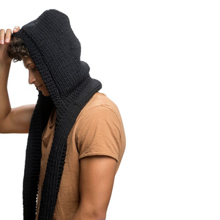 A person in a tan shirt adjusting a large knitted black Champion Hood Scarf made of merino wool with sherpa fleece lining, looking down and to the side, against a white background.
