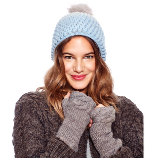 A woman with a warm smile wearing a blue knit hat with a pom-pom, Double Cable Knit Handwarmers, and a textured dark sweater.